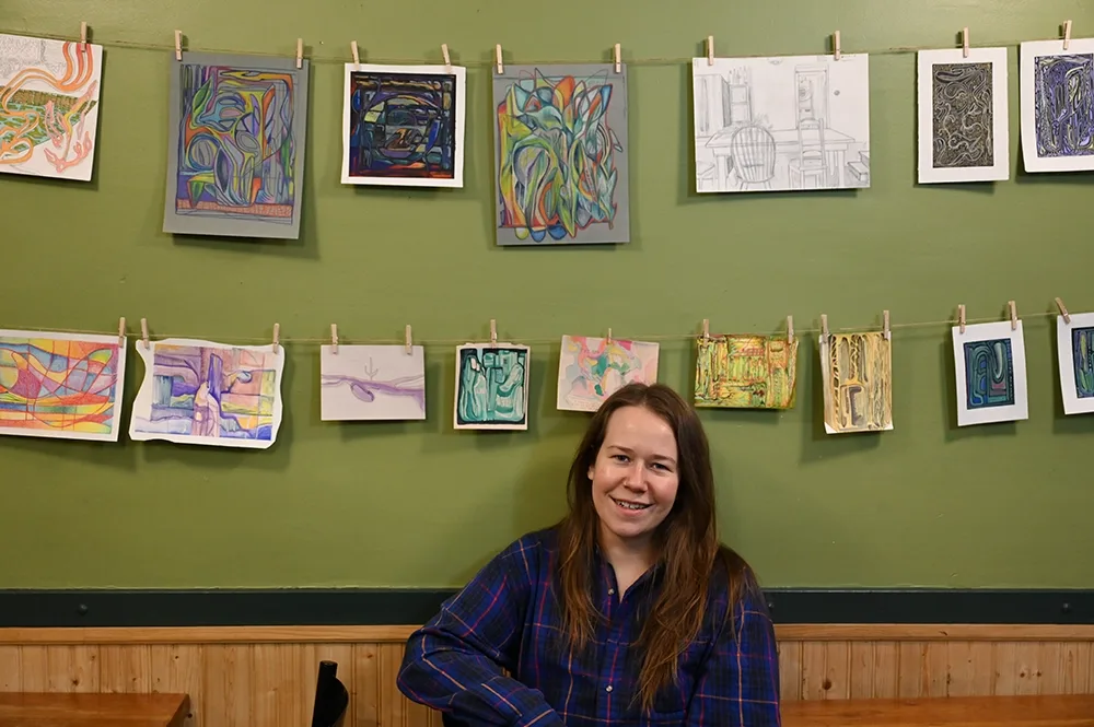 A person sits in front of their artwork. The artwork is around 15 sketches and paintings, in two rows, hung from clothes pins on a string, like laundry.