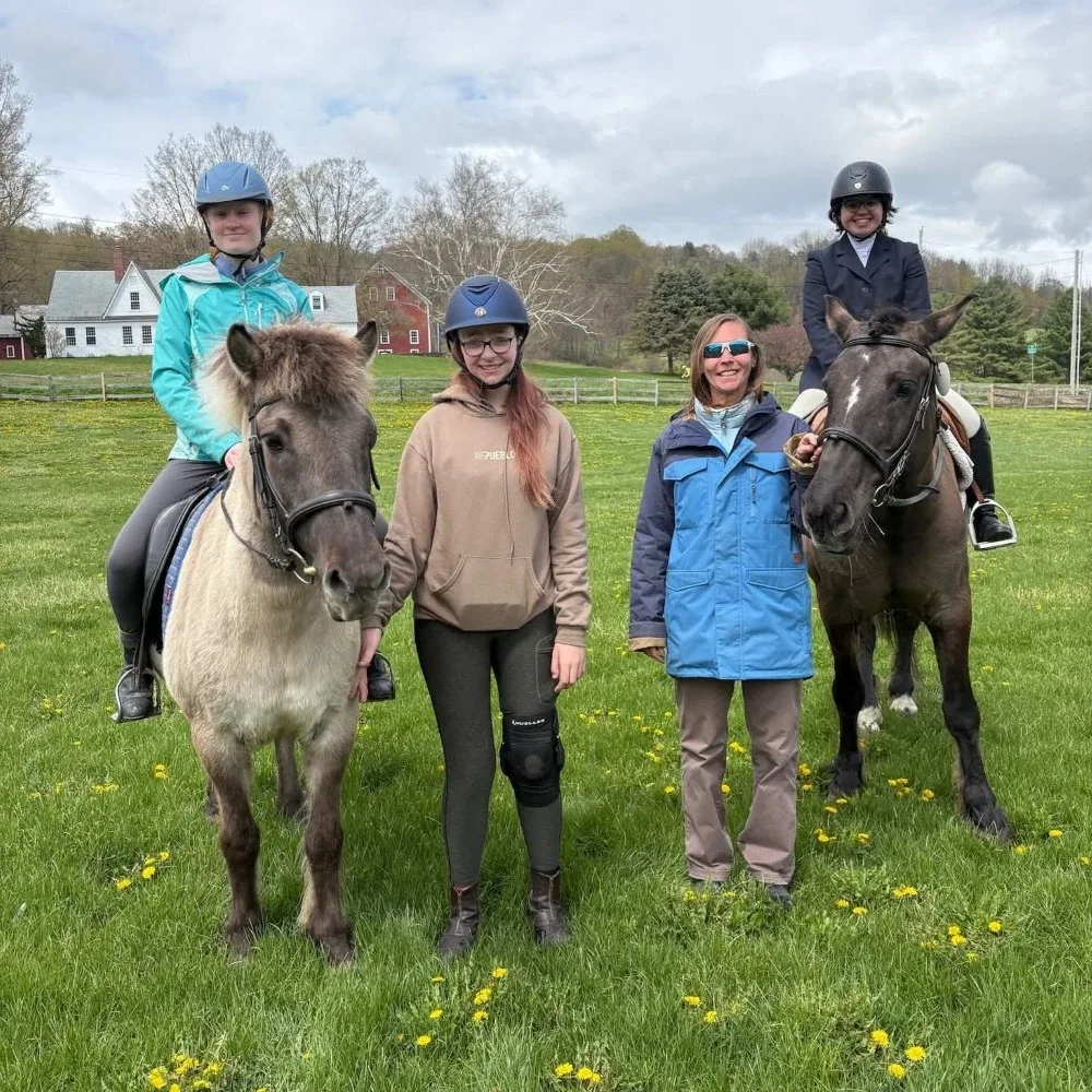 From left to right, Charlotte Graham, Olivia Arnell, Katherine Wagenbach '98, and Logan Lipschultz pose for a photo in a field. Charlotte and Logan are on horseback, while Olivia and Katherine are standing.