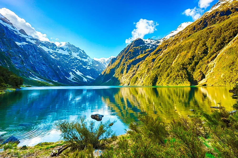 Lake Marian New Zealand on a sunny day with blue water and rocky green mountains that have snowy faces.