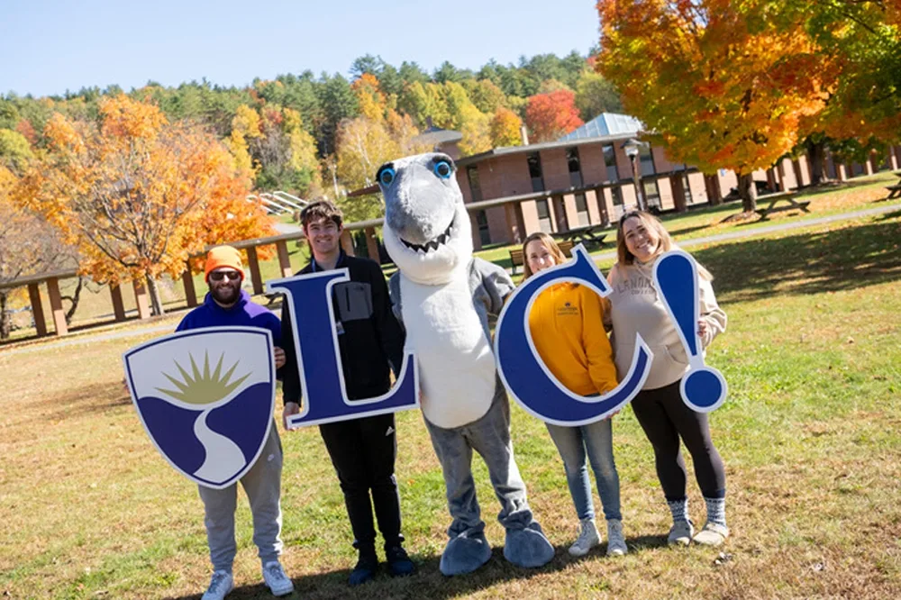 Four students holding cut outs including the college shield, letters L and C, an exclamation point, with Finn the Mascot on the quad on a sunny autumn day.