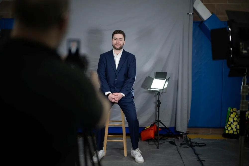 A student sits on a stool in front of a gray backdrop while a headshot is taken for their LinkedIn profile.