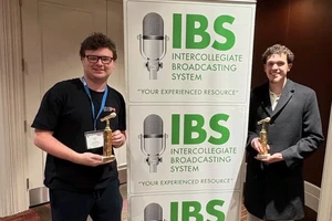 Two students holding their award trophies while standing next to an Intercollegiate Broadcasting System pull-up banner.