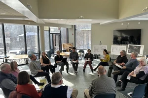 A group of 15 people sitting in a circle and having a conversation. Behind them are three easels with floor plans of buildings and aerial photography of campus