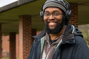 Photo of Anastasio Bonhomme smiling for camera on Landmark College quad. He is wearing a wool hat and headphones.