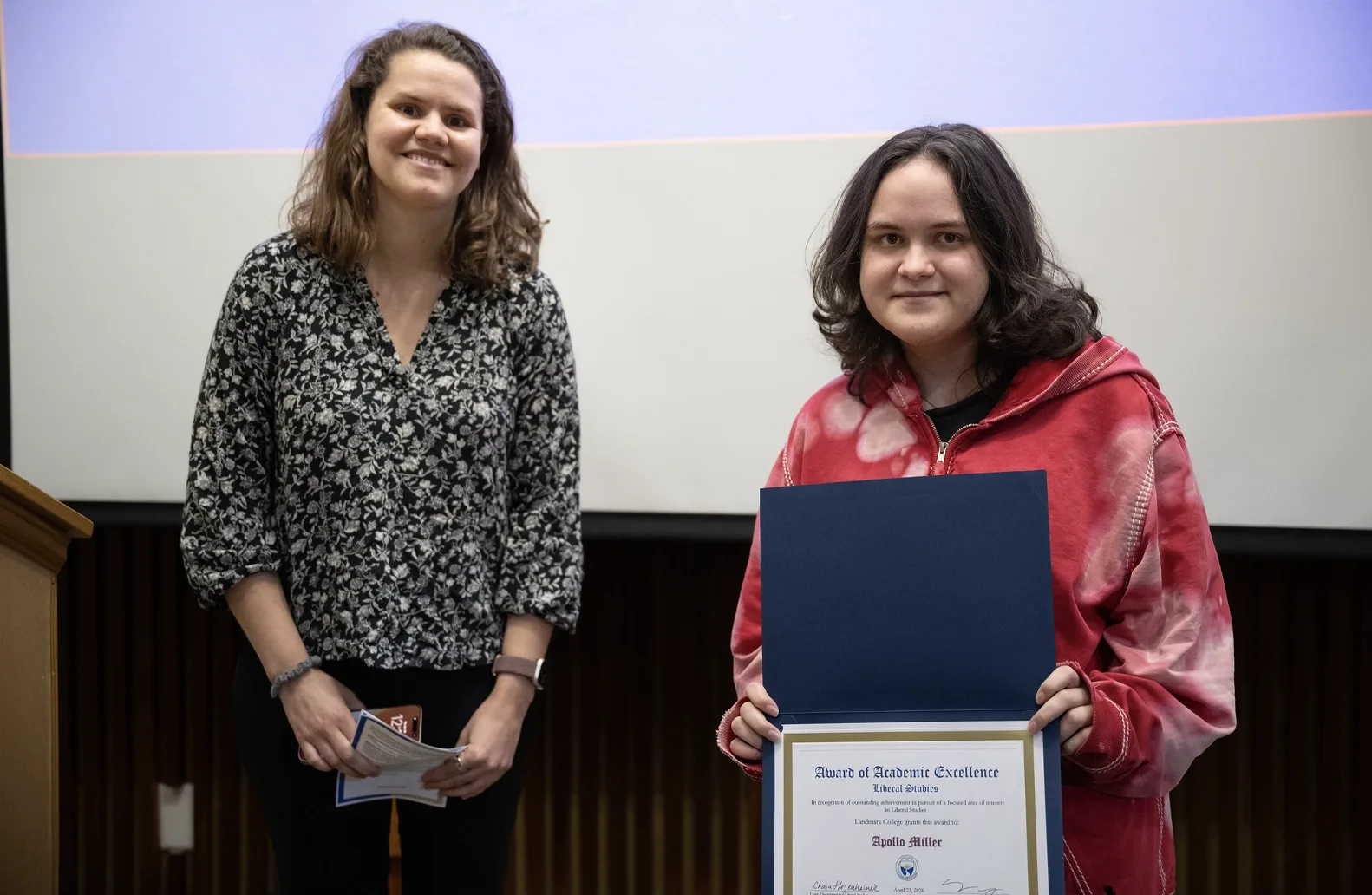 A professor and a student stand in front of a screen at an academic awards event. The student, right, holds an award displaying the student's name.