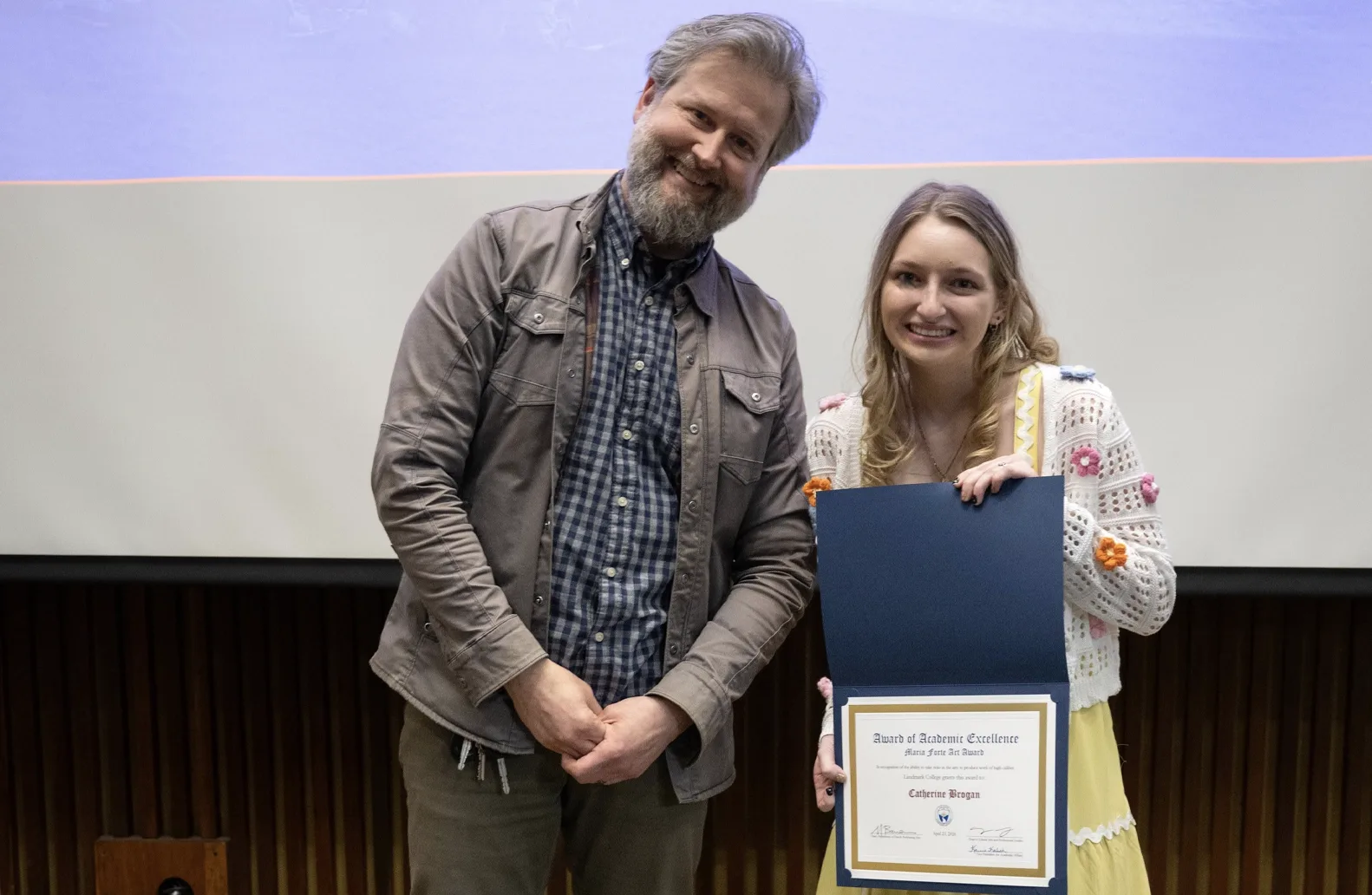 A professor and a student smile and stand in front of a screen at an academic awards event. One of the individuals, a student wearing a white sweater with colorful flowers, right, holds an award displaying her name.