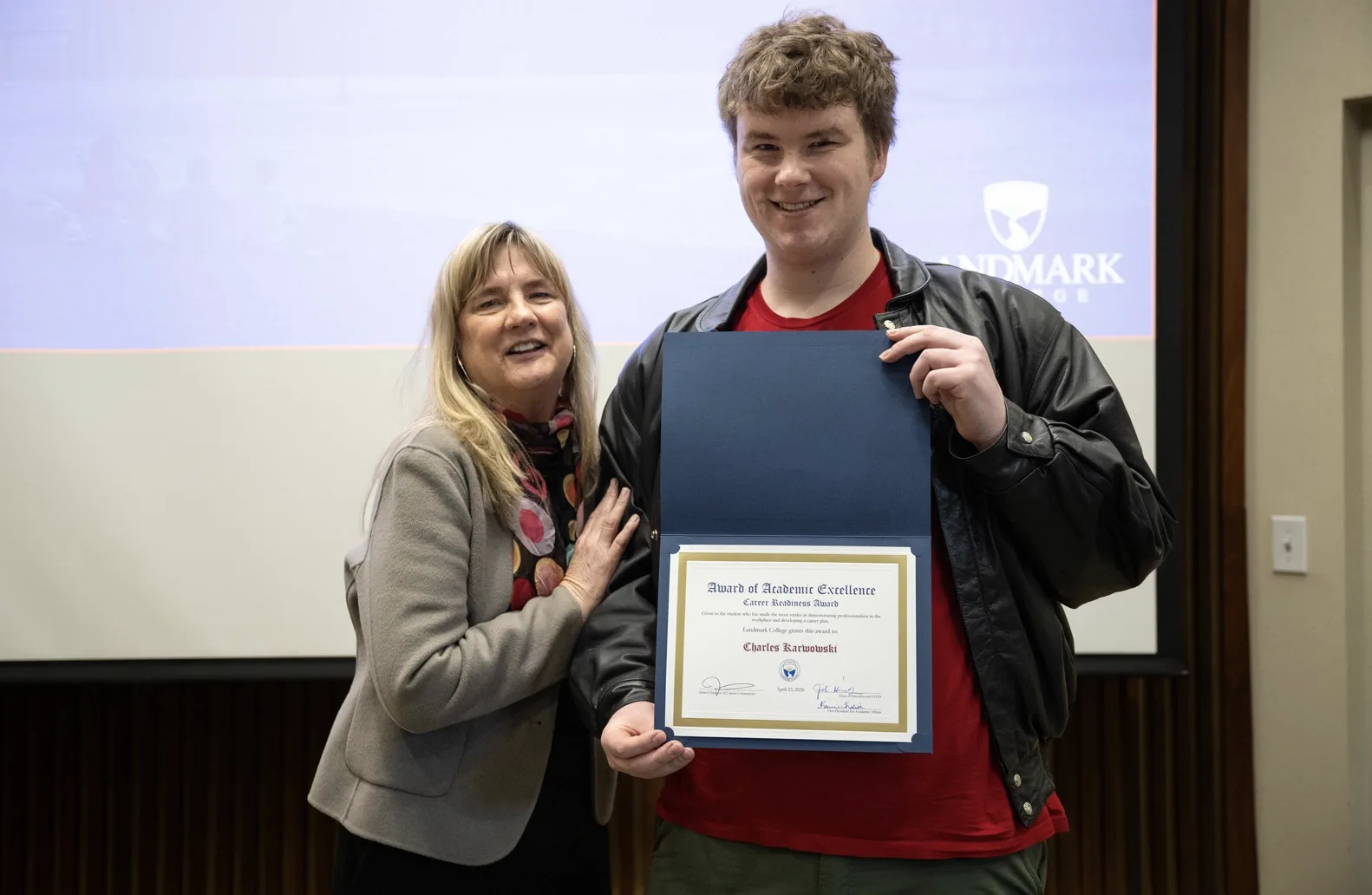 Two people stand in front of a screen at an academic awards event. The woman on the left has her hand on the student on the right's arm. The student, wearing a red shirt and leather jacket, holds an award displaying the student's name.