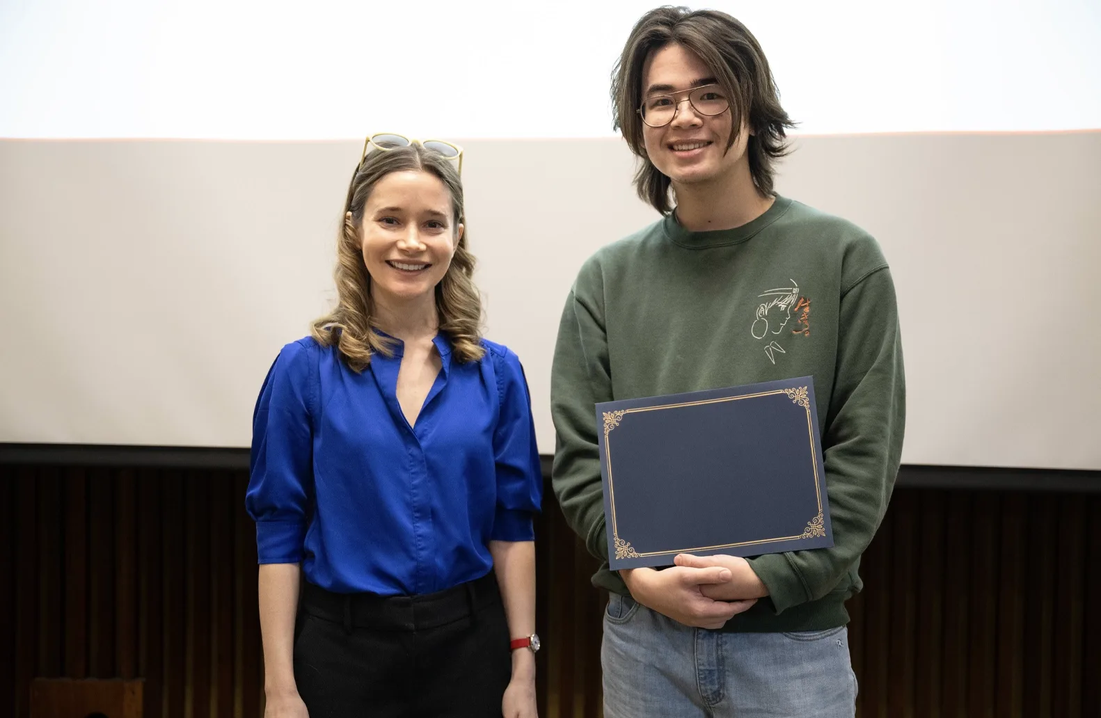 A professor in a blue shirt and a student in a green long-sleeved shirt smile and stand in front of a screen at an academic awards event. The student, right, holds an award.
