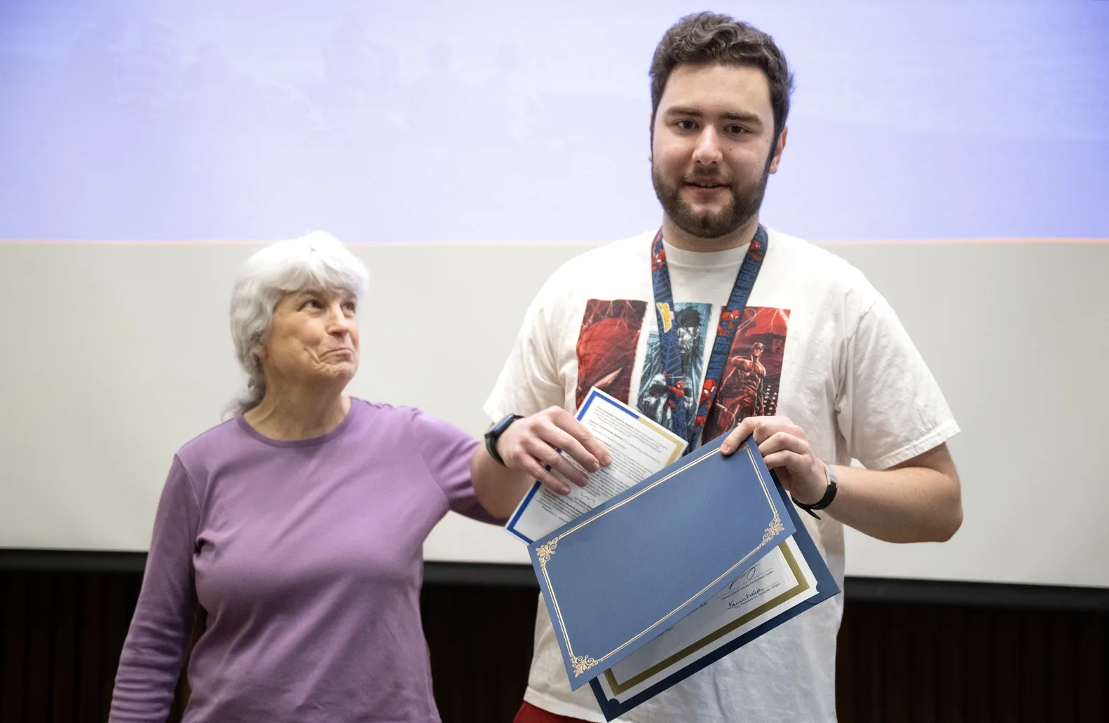 Two people stand in front of a screen at an academic awards event. One of the individuals, a student, right, holds an award.