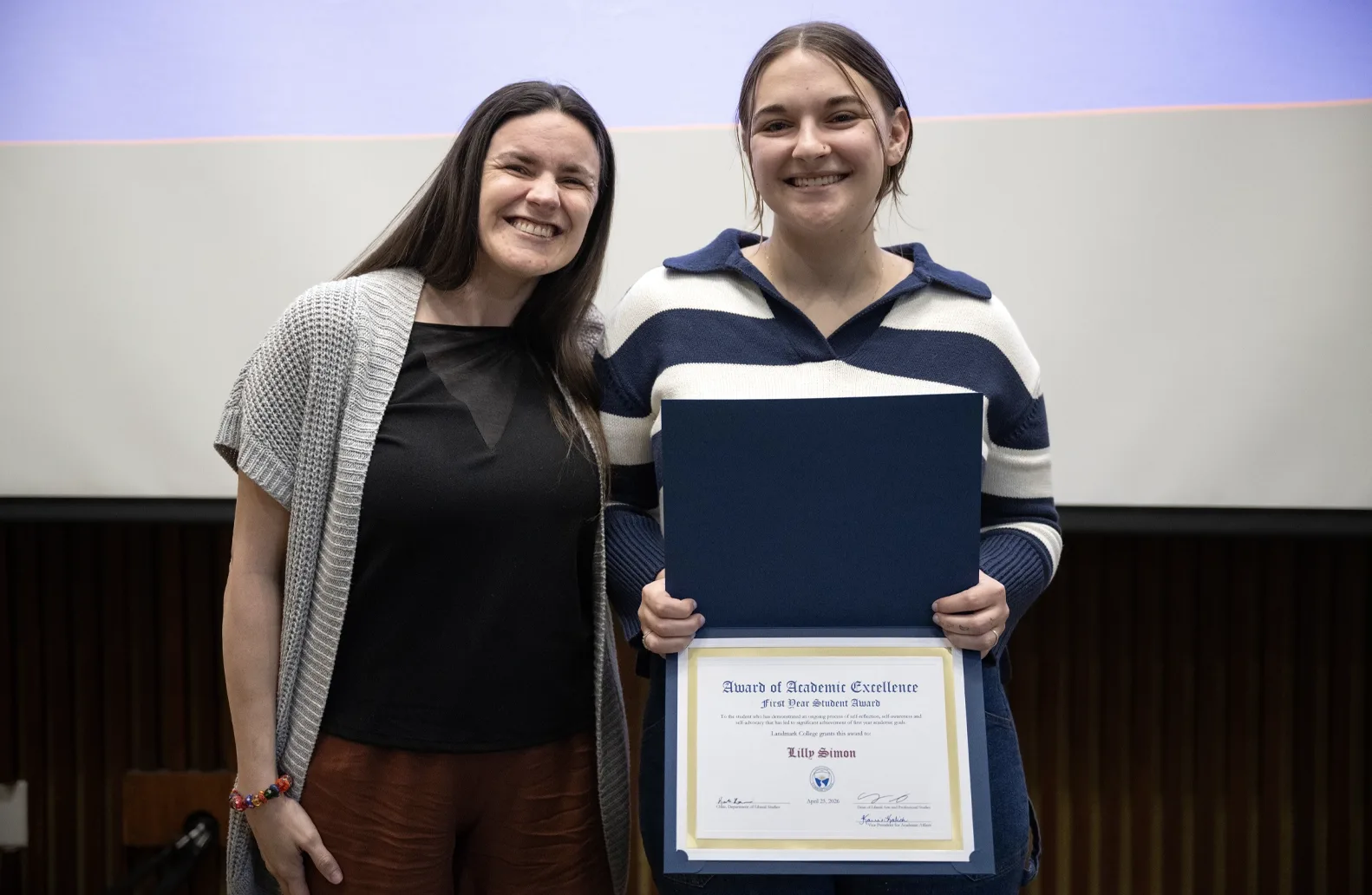 Two people stand in front of a screen at an academic awards event. One of the individuals, a student wearing stripes, right, holds an award with the student's name displayed.