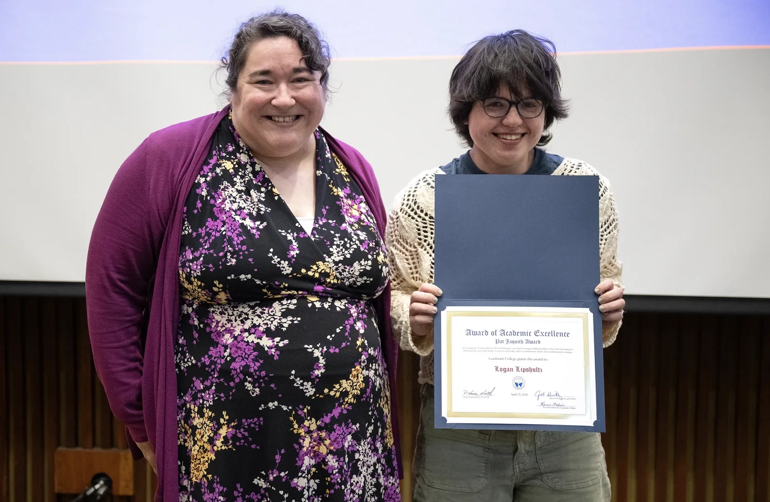 Two people stand in front of a screen at an academic awards event. One of the individuals, a student, right, holds an award.