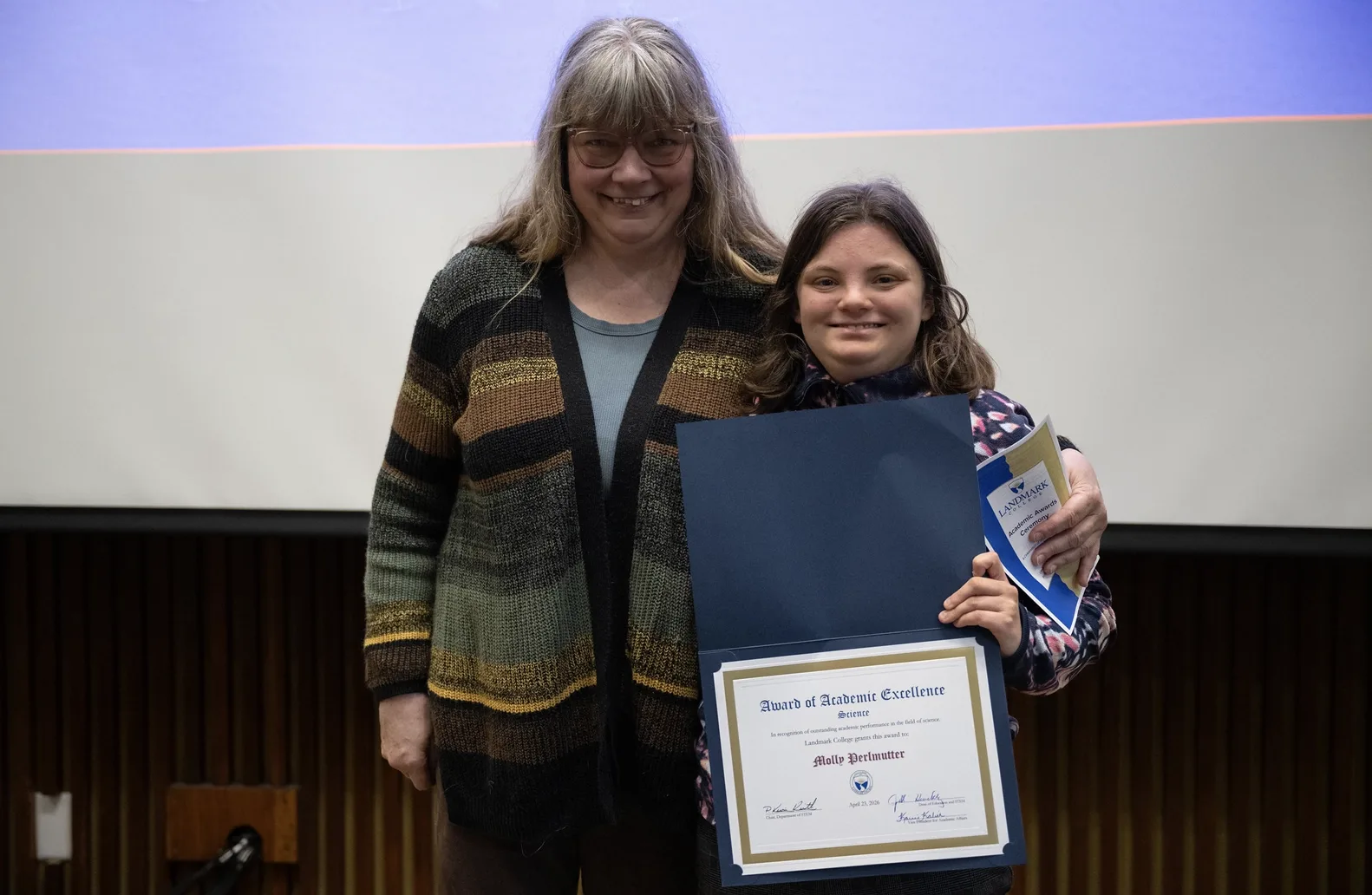 Two people stand in front of a screen at an academic awards event. One of the individuals, a student, right, holds an award displaying the student's name.