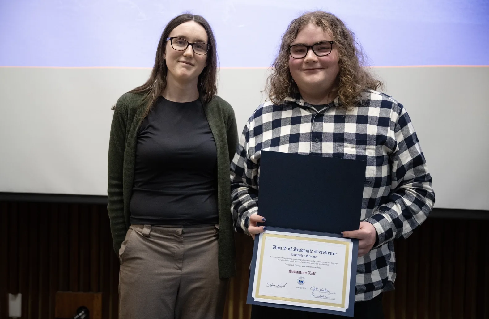A professor and a student smile while standing next to each other. The student, in a black and white checkered shirt, is holding an award displaying the student's name.