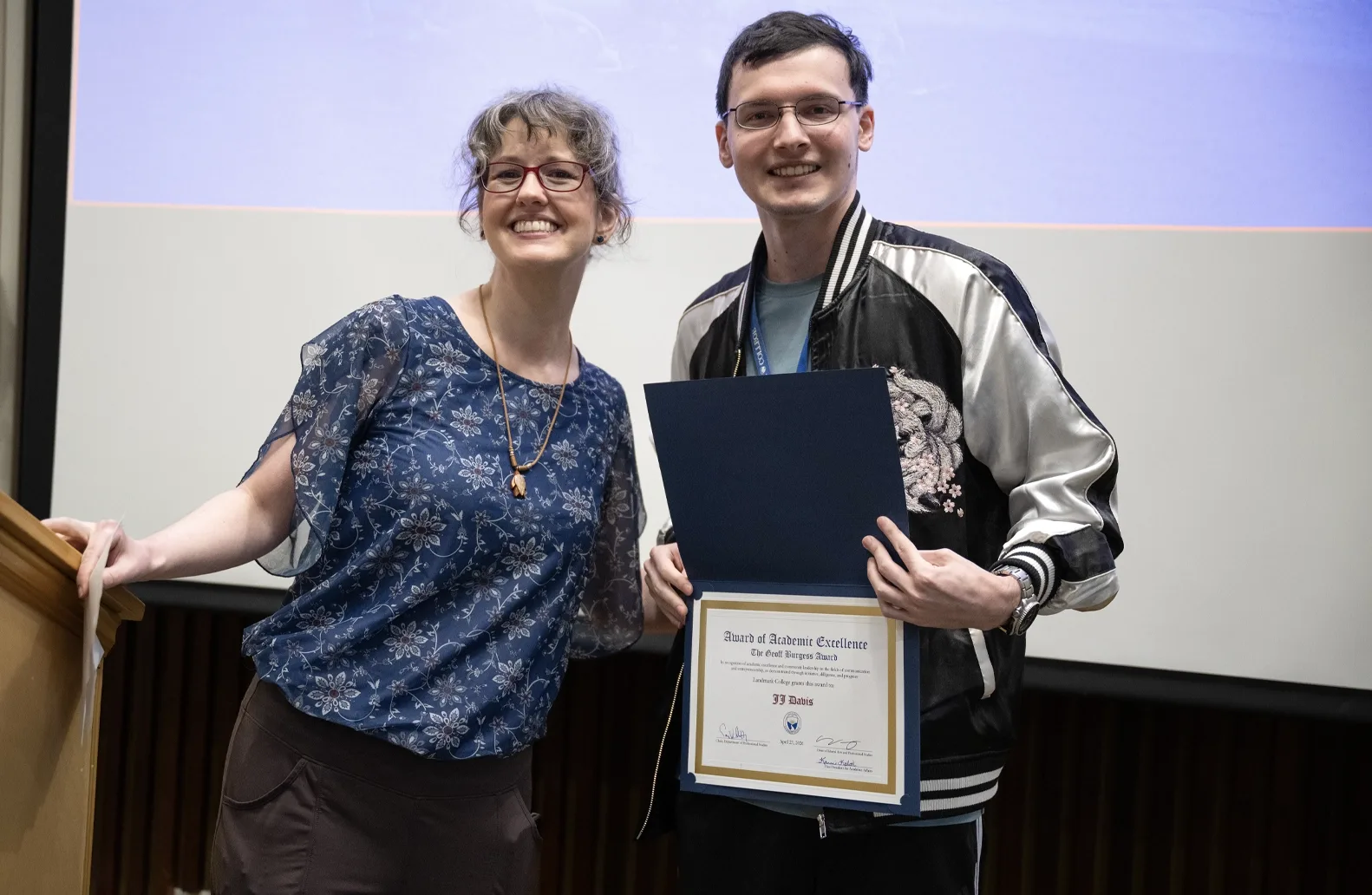 Two people stand in front of a screen at an academic awards event. One of the individuals, a student, right, holds an award displaying the student's name.