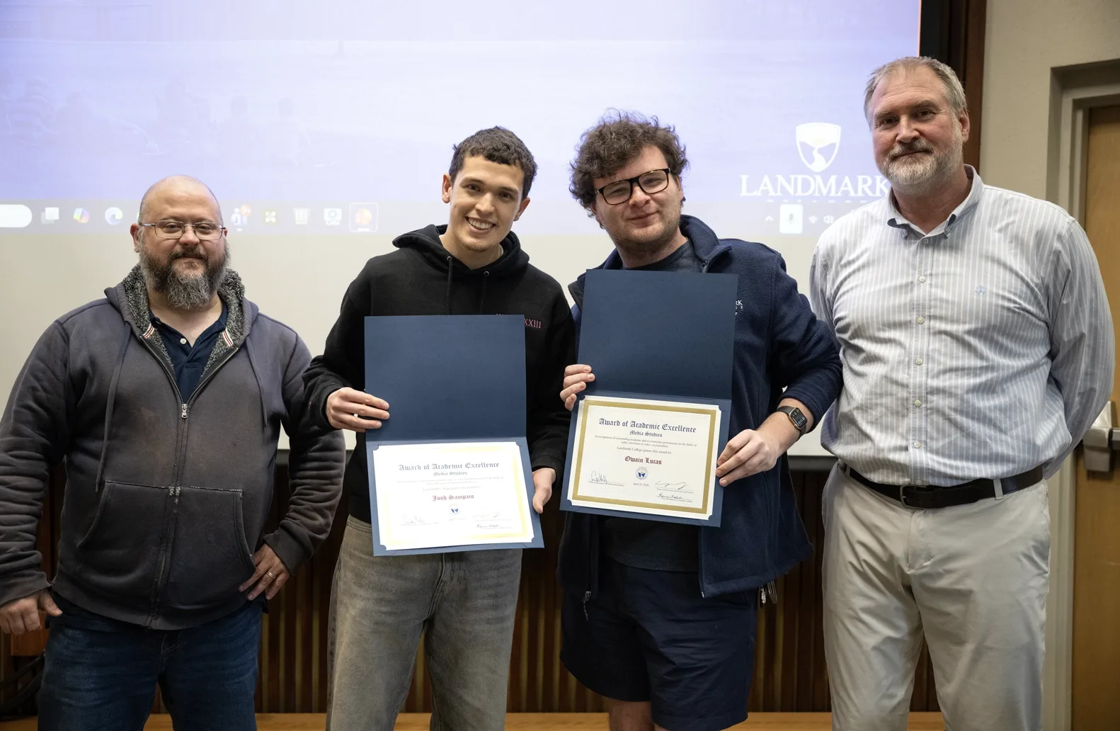 Four people stand in a row at an academic awards ceremony. The students in the middle hold awards displaying their names.