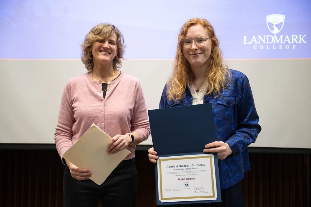 A professor, in light pink, holds some papers. At right is a student, in blue, who has been presented with an award. The students holds up the award so her name is displayed. On a screen behind the student's shoulder is the Landmark College logo.