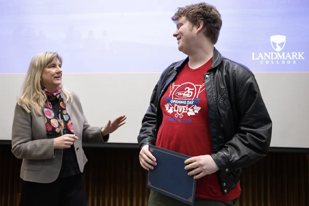 A student in a red shirt and leather jacket, right, holds an award. A staff member in a colorful scarf and neutral blazer, left, speaks. On a screen behind the student's shoulder is the Landmark College logo.