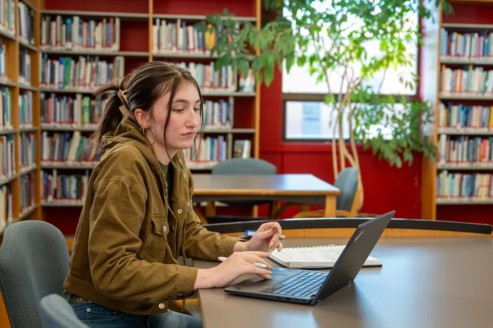 Student sitting at a table in a library, and typing on a laptop.