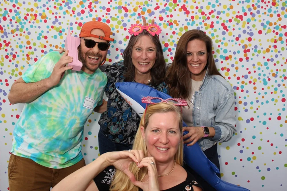Four people smiling and posing with novelty accessories against a multicolored polka dot backdrop.