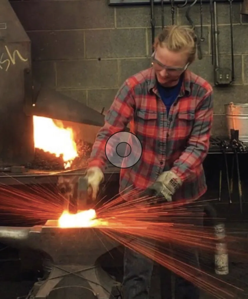 Person blacksmithing in a workshop, striking hot metal on an anvil, causing orange sparks to fly.