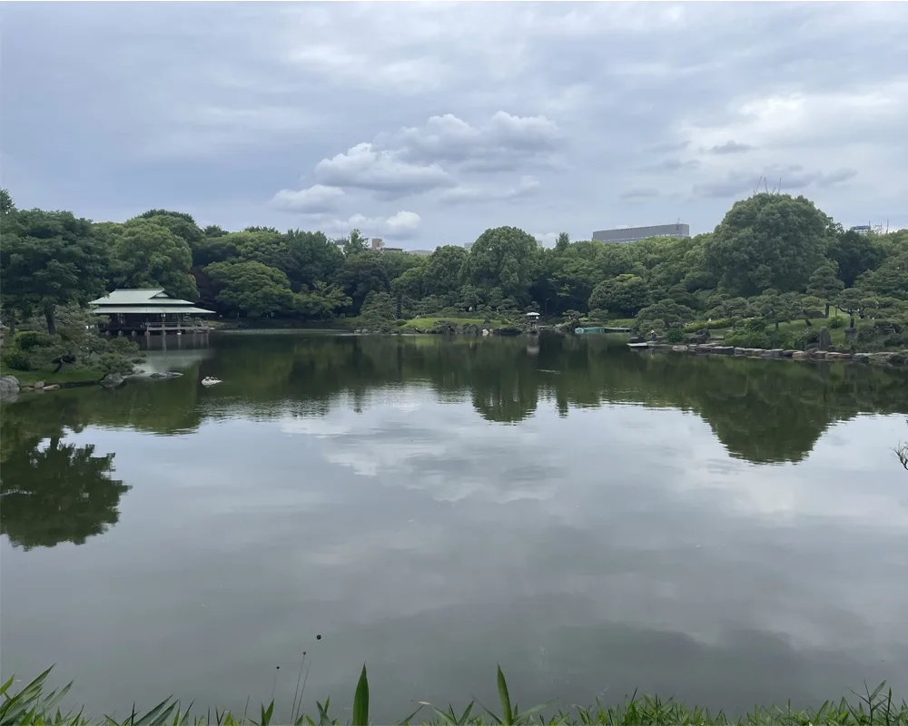 A large, still pond surrounded by trees with a Japanese tea house perched on the edge of the water. The trees and cloudy sky are reflected in the pond.