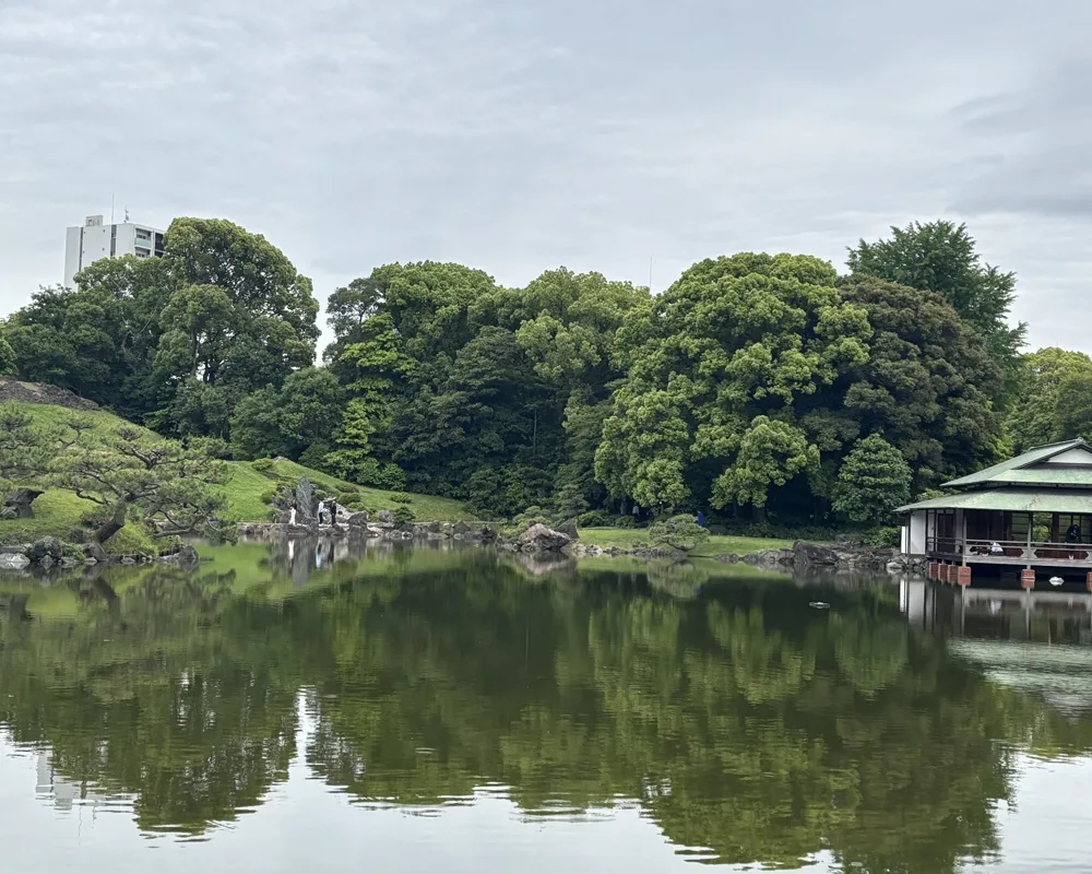 A serene Japanese garden with a pond reflecting lush green trees, a traditional tea house on the right, and a modern building peeking through the foliage in the background.