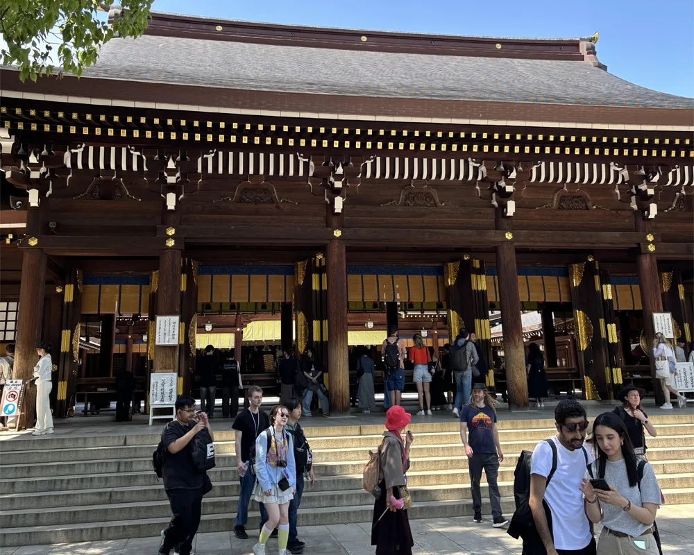 Visitors gather outside a traditional wooden Shinto shrine under a clear blue sky, with green tree branches framing the top left corner.