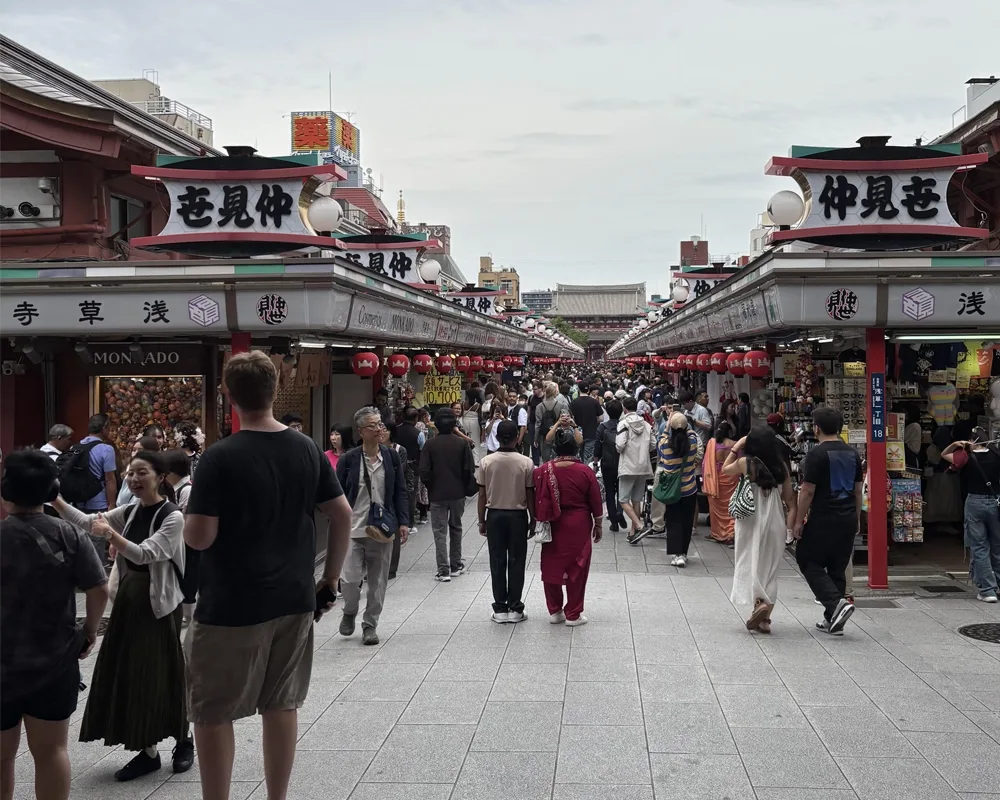 A bustling shopping street lined with traditional-style stalls and lanterns, filled with people walking toward a temple in the distance under an overcast sky.