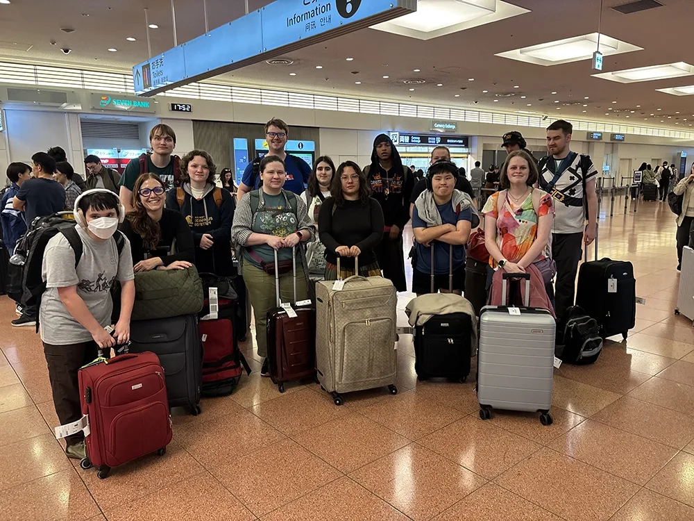A group of student travelers and a faculty member with luggage pose for a photo in a Japanese airport, looking tired but happy.