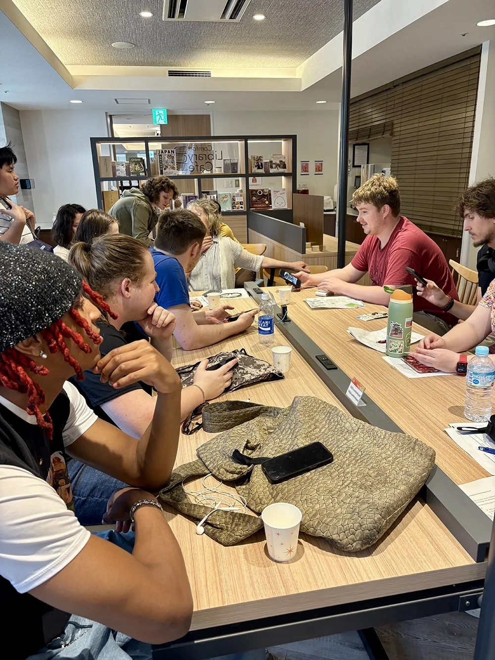 A group of young people are sitting and standing near tables, are looking at their phones, and looking at each other.