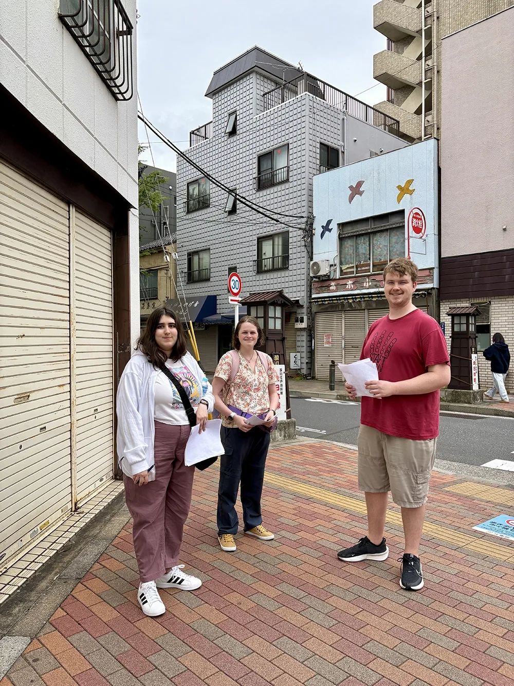 Three people are standing on a street corner; each person is holding pieces of paper.