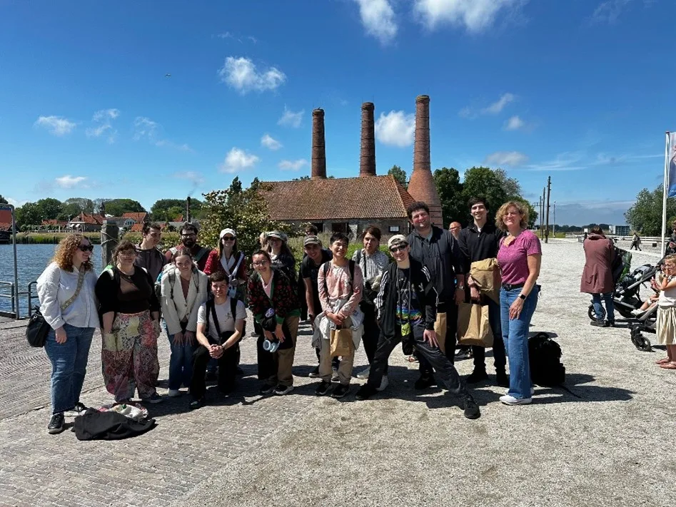 A group of 16 smiling people stand in bright sunlight with water, a building and three brick smokestacks in the background.
