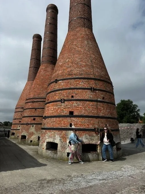 Two students stand in front of three large brick structures with ovens at the base tapering into tall smokestacks