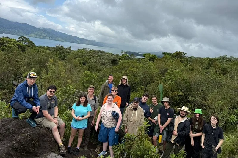 Students and trip leaders pose for a photo on rock with a view of a lake and mountains in the distance.