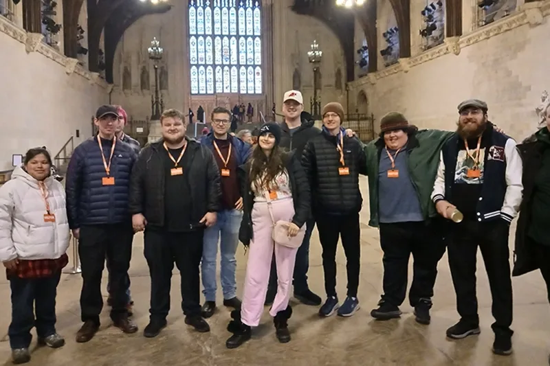 A group of young men and women stand in a medieval great hall with a vaulted ceiling and exposed wood beams. There is a large stained glass window in the background.