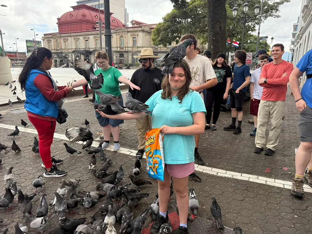 A female student holding a bag of Cheetos is surrounded by pigeons as well as one perched atop her head. Other students in the background are also interacting with the pigeons.