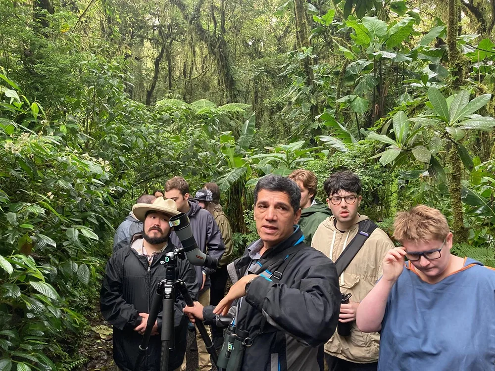 The Manuel Antonio National Forest tour guide stands at the front of a group of students holding a camera on a tripod talking about the lush plant life surrounding them.