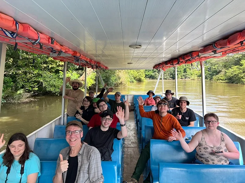 A large group of individuals wave at the camera while seated on a long, narrow boat with a canopy roof.