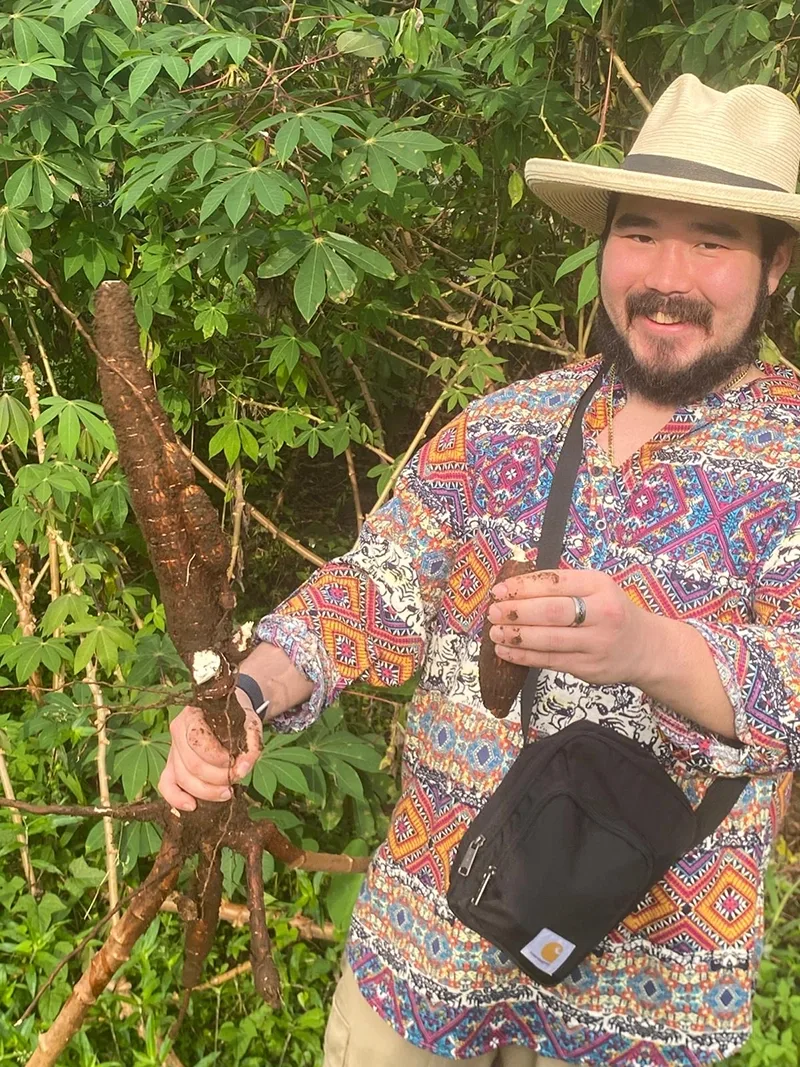 Student with dark mustache and beard wearing white wide-brimmed hat and a bright patterned shirt smiles at the camera while holding up a long brown root.