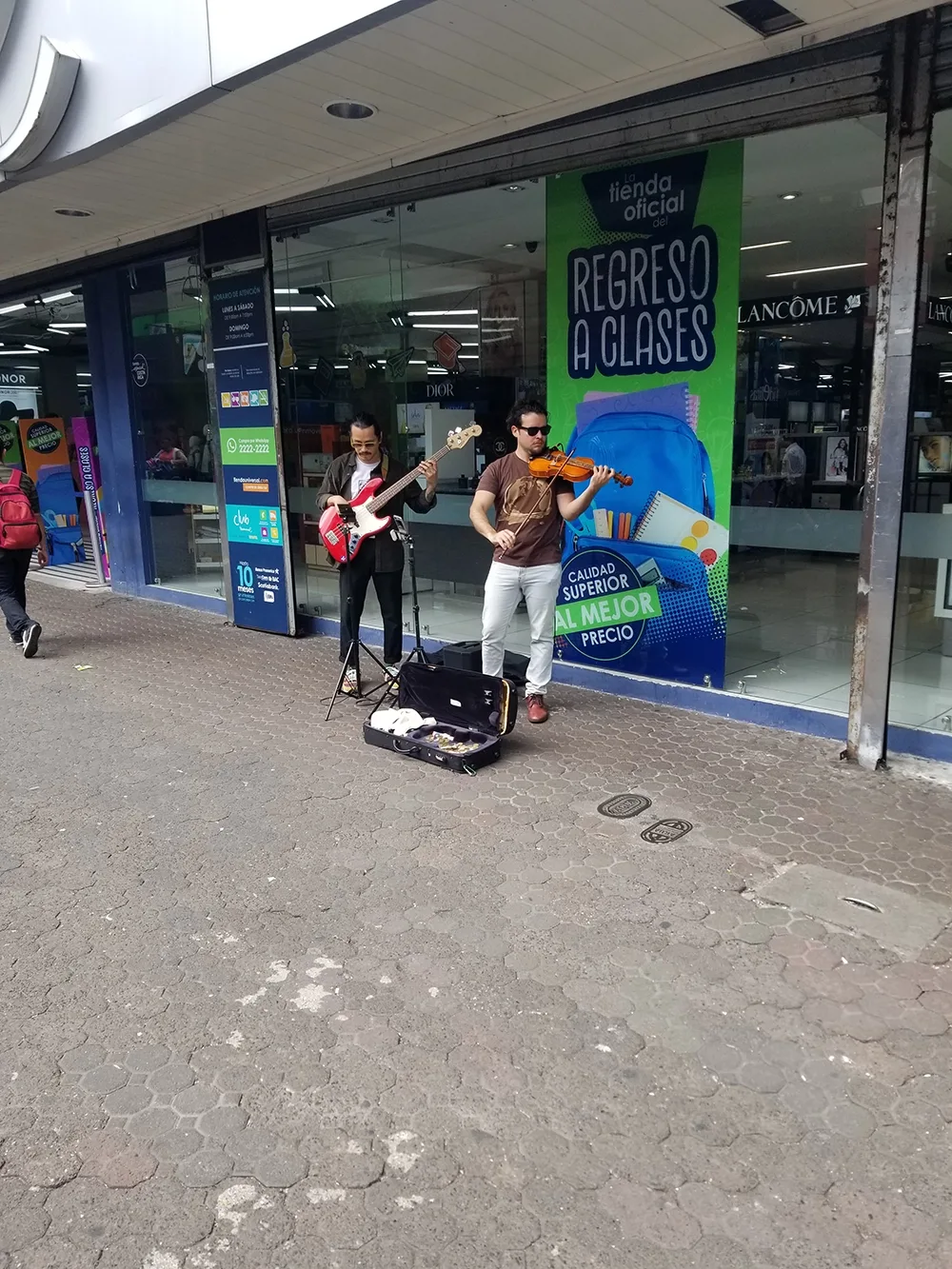 An electric bass player and a violinist perform in front of a storefront with tall glass windows and banners in Spanish.