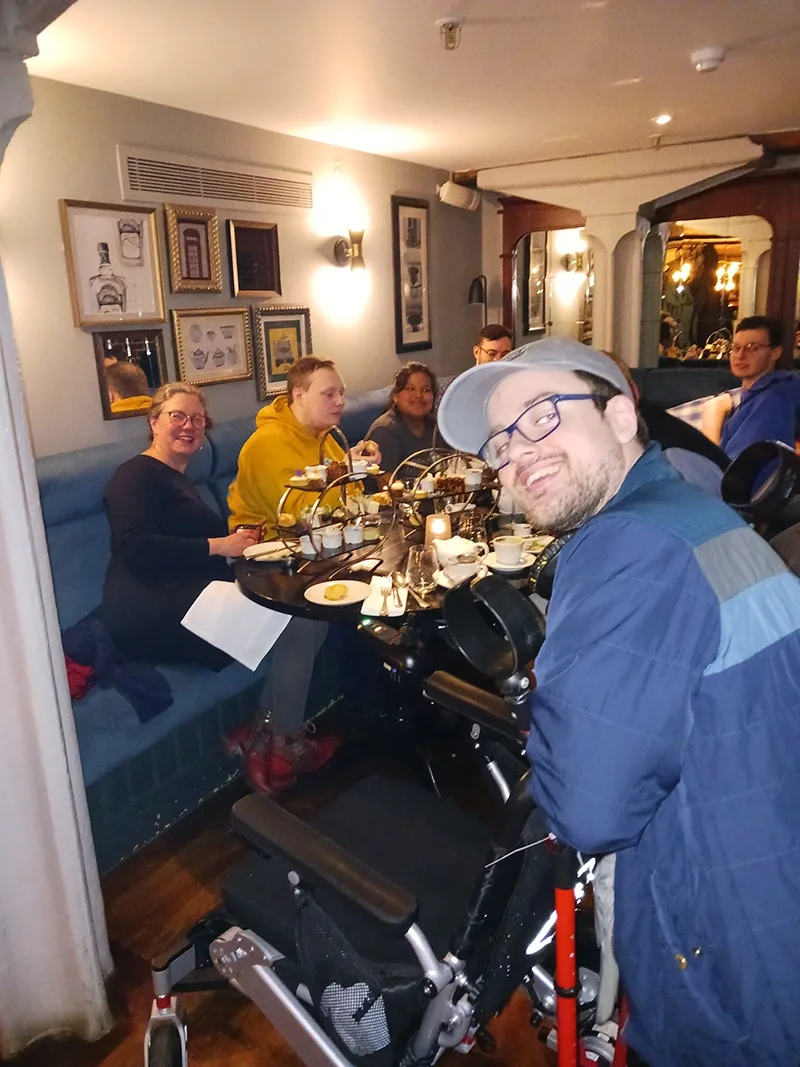 Several young men and women appear seated at a table in a restaurant with a traditional English afternoon tea spread in front of them.