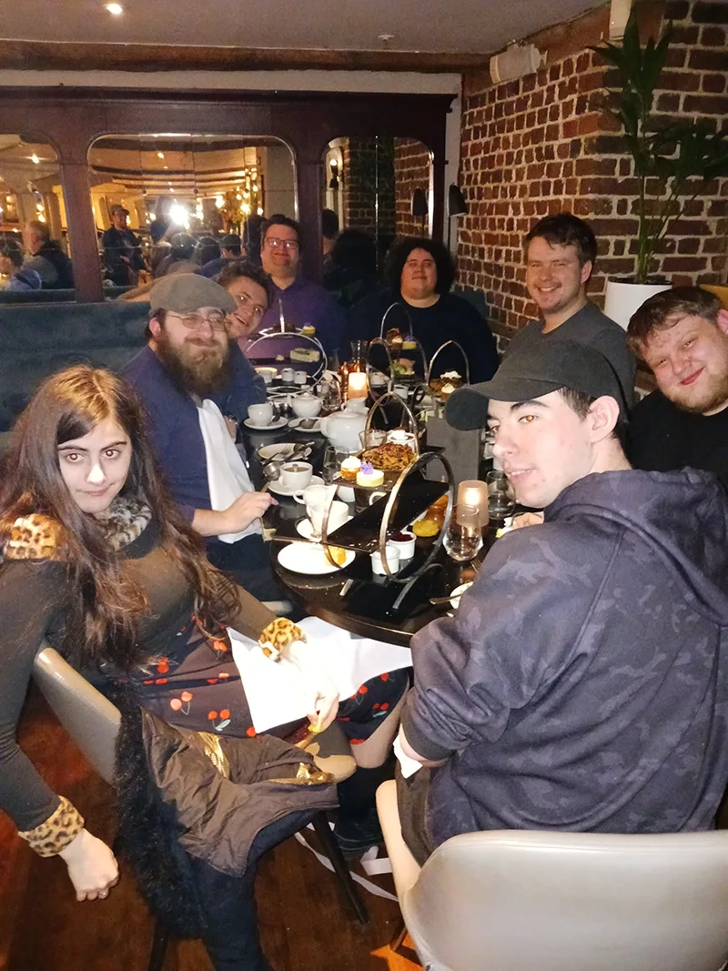 Several young men and one young woman appear seated at a restaurant dining table with a traditional English afternoon tea spread in front of them.