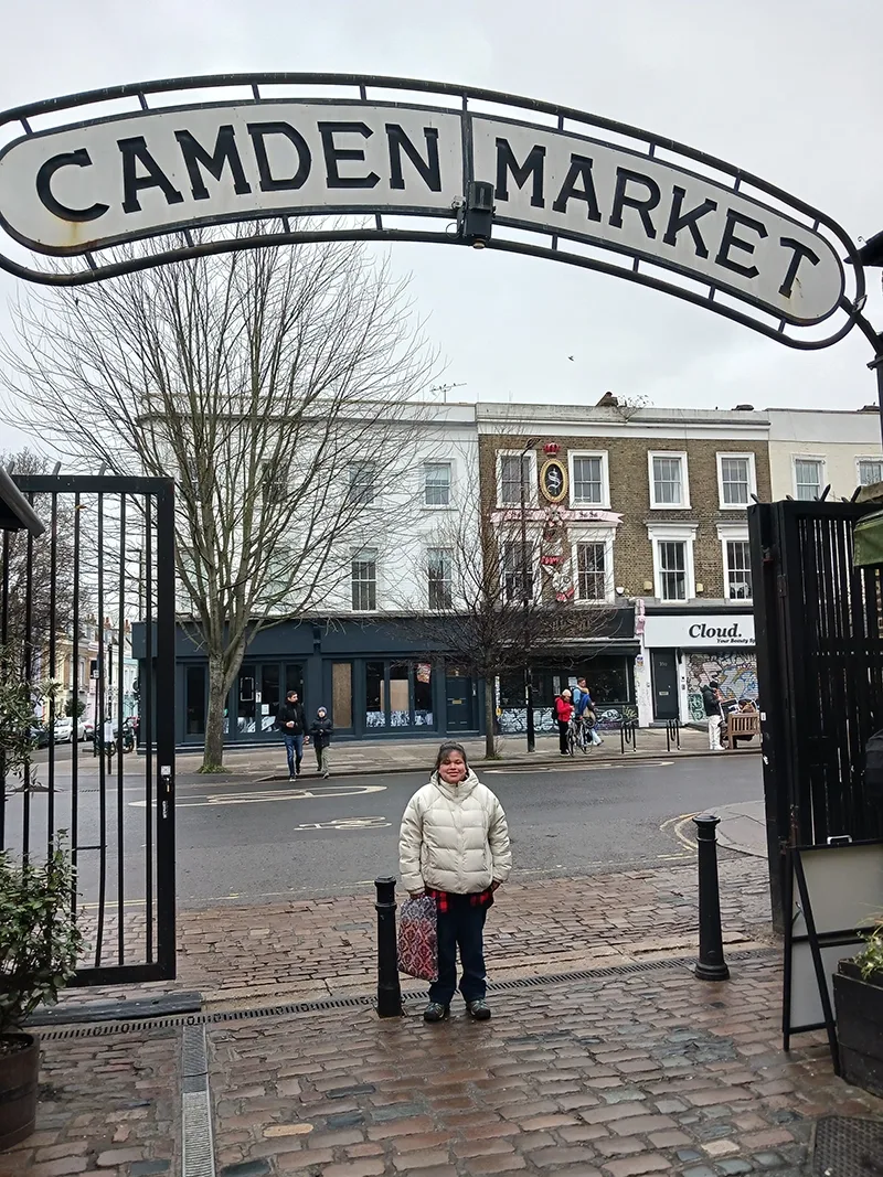 A young woman in a white winter coat stands beneath an arched metal Camden Market sign.