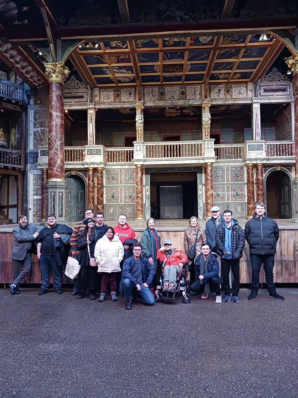 A group of young adult students and their instructors stand in front of a large stage at the Globe open air theater in central London.