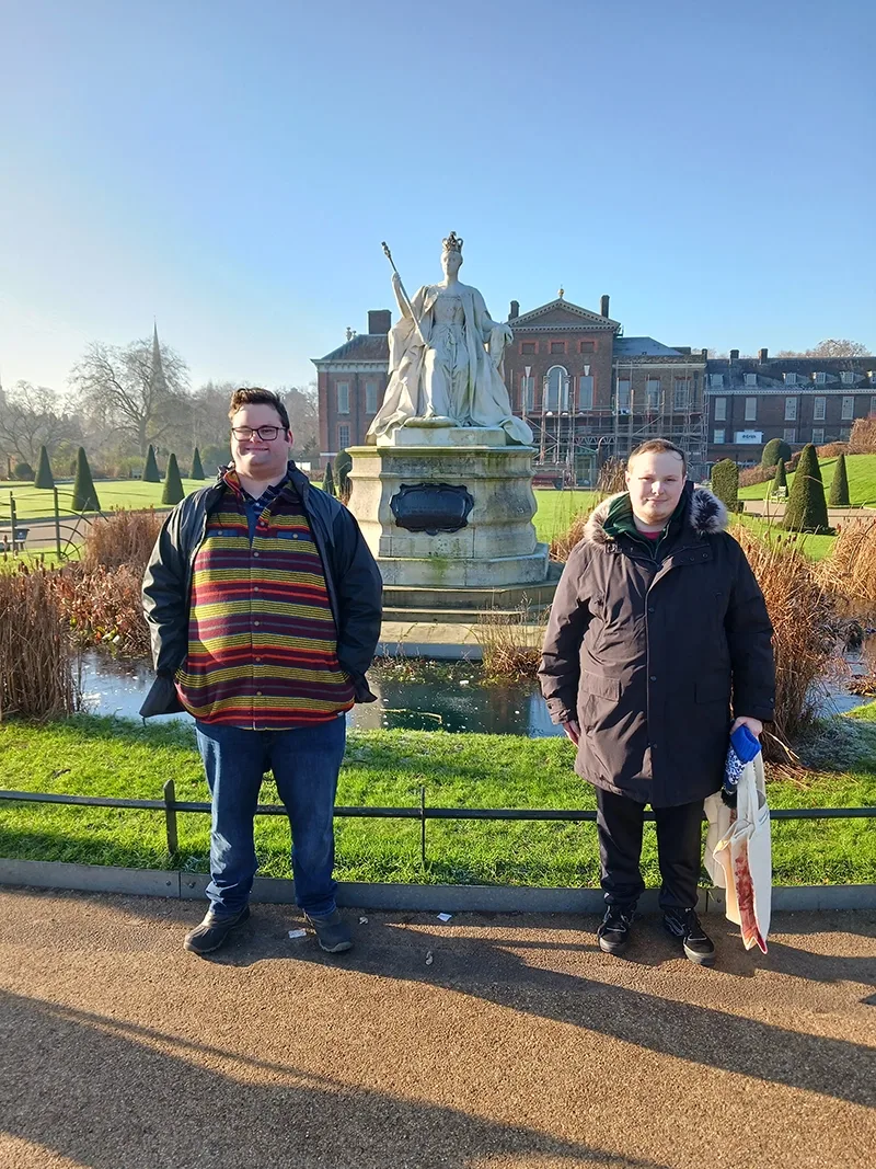 Two young men in winter jackets stand in Hyde park in front of a statue of queen Victoria with Kensington palace in the background.