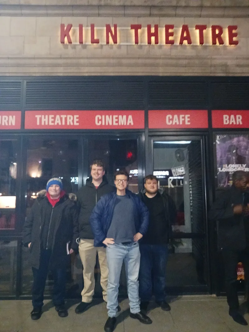 Four young men in winter jackets stand in front of a theater building at night in north London.