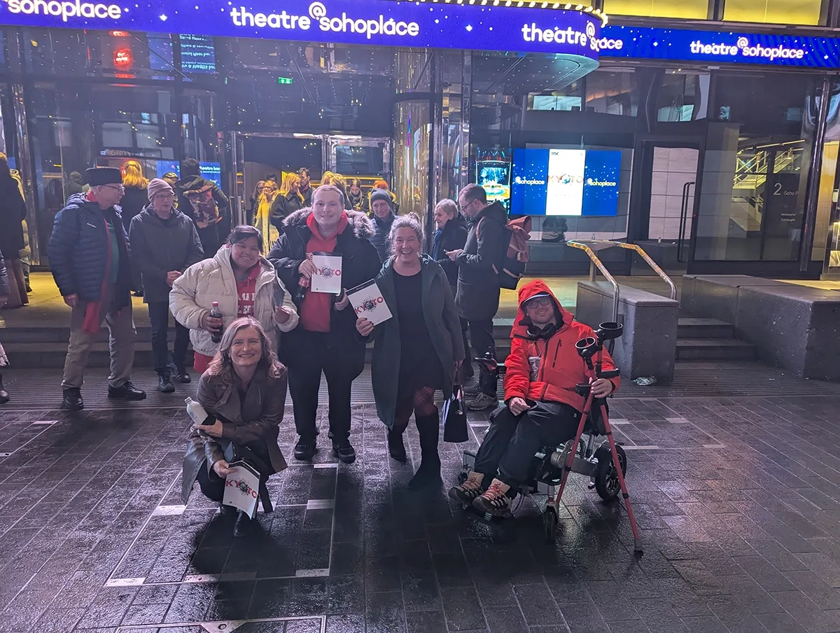 Five men and women (including a young man in a wheelchair) stand in front of a theater building in central at night holding program publications for a theater performance of Kyoto.
