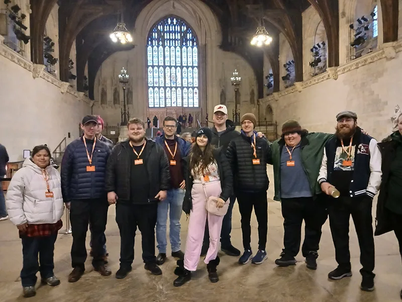 A group of young men and women stand in a medieval great hall with a vaulted ceiling and exposed wood beams. There is a large stained glass window in the background.