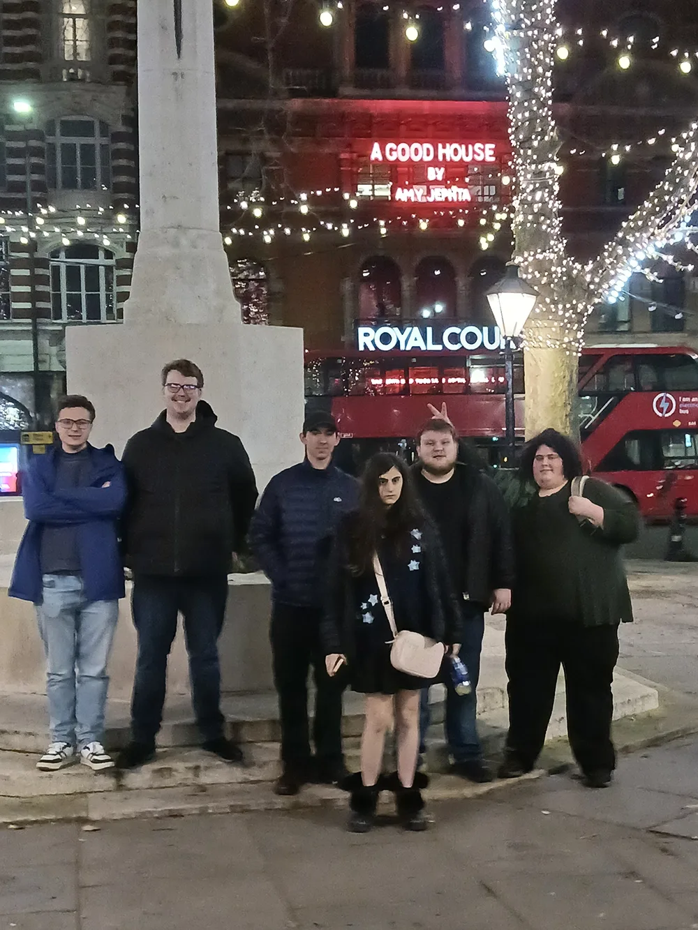 Five young men and one young woman stand in Soho Square in central London at night with a lighted theater marquee in the background.