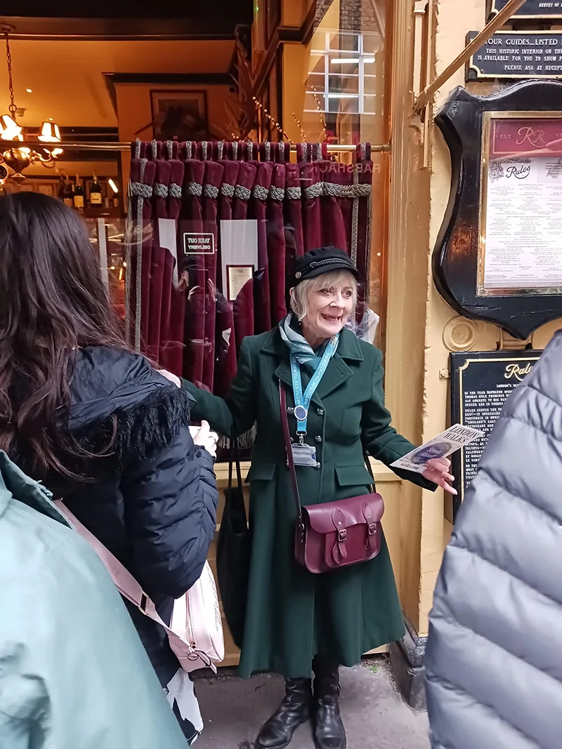 A woman stands in a long green coat and black hat outside a restaurant building in central London